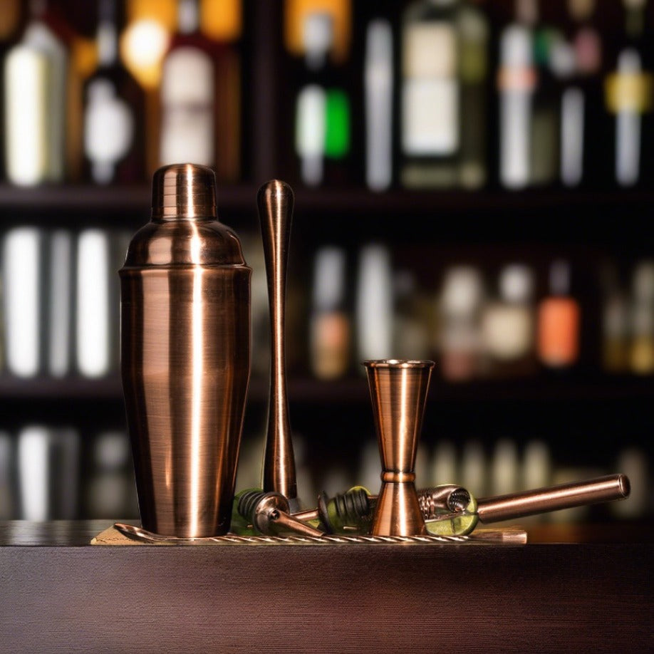 A set of brushed copper cocktail shaker, muddler, bar spoon, strainer, pourers, and a double jigger arranged on a wooden surface with a blurred background of bottles.
