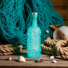Blue rusty barrel spirits gin bottle with silver cap on a wooden surface with fishing net and ropes in the background