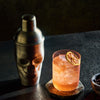 Rusty Barrel Spirits Skull-shaped cocktail shaker next to a glass of orange cocktail with a lemon wheel on a dark surface.
