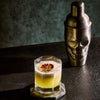 Cocktail in a glass with a skull-shaped shaker on a dark surface