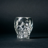 A clear shot glass shaped like a skull, placed against a dark background.