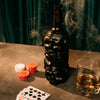 Skull-shaped bottle of Rusty Barrel Caribbean Gold Rum with smoking effect, poker chips, playing cards, and a glass of whiskey on a table.