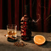 Skull-shaped bottle of Rusty Barrel Spiced Rum with a glass, orange, and cigars on a dark surface.