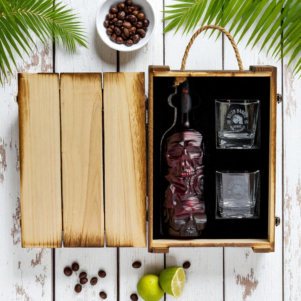 Rusty Barrel Spiced Rum bottle and two glasses in a wooden box with coffee beans and limes on a rustic surface.