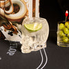 Clear skull-shaped glass with a lime wedge on a black surface with playing cards and a cocktail in the background.