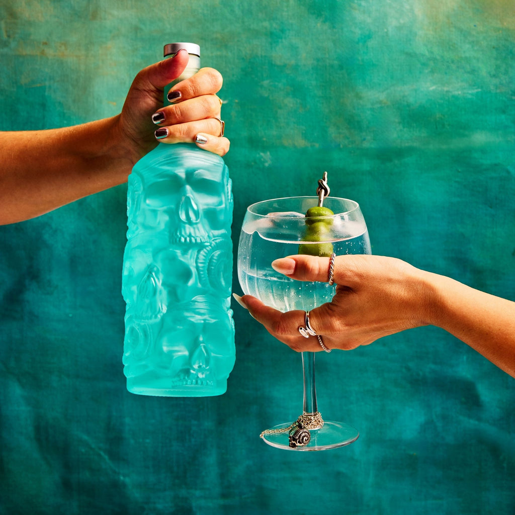 Person holding a blue bottle of Rusty Barrel London Dry Gin and a glass with a cocktail against a teal background