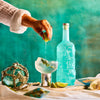 Person preparing a cocktail with a blue bottle of Rusty Barrel London Dry Gin and lime garnish against a teal background