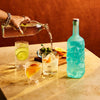 Person pouring mixer into a glass with a blue bottle of Rusty Barrel London Dry Gin on a table.