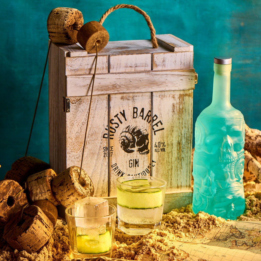Rusty Barrel London Dry Gin bottle and glasses with a rustic wooden crate on a textured surface.