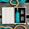 Rusty Barrel London Dry Gin Teal bottle and glasses in a white wooden box with fishing net and rope on a wooden surface