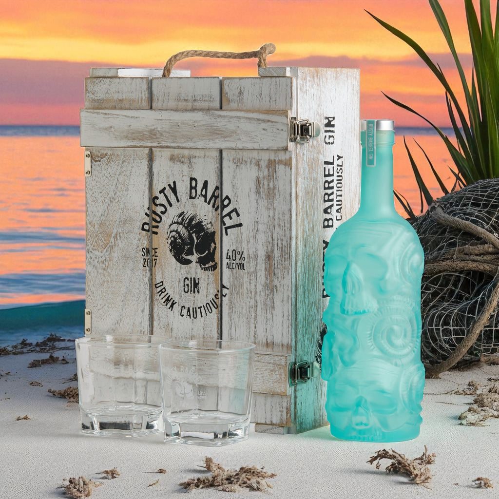 Teal Rusty Barrel London Dry Gin with decorative design, wooden crate labeled 'Rusty Barrel Gin', and glasses on a beach with sunset background.
