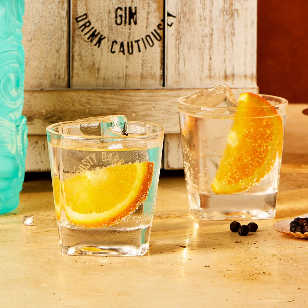 Two glasses of Rusty Barrel gin with orange slices on a wooden surface, with a gift box in the background.