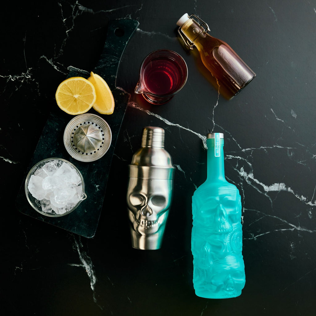 Cocktail-making setup with a skull-shaped shaker, Rusty Barrel Gin bottle, glass with ice, and lemon slices on a dark surface.