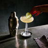 Cocktail with a lime garnish on a bar counter next to a skull shaped cocktail shaker.