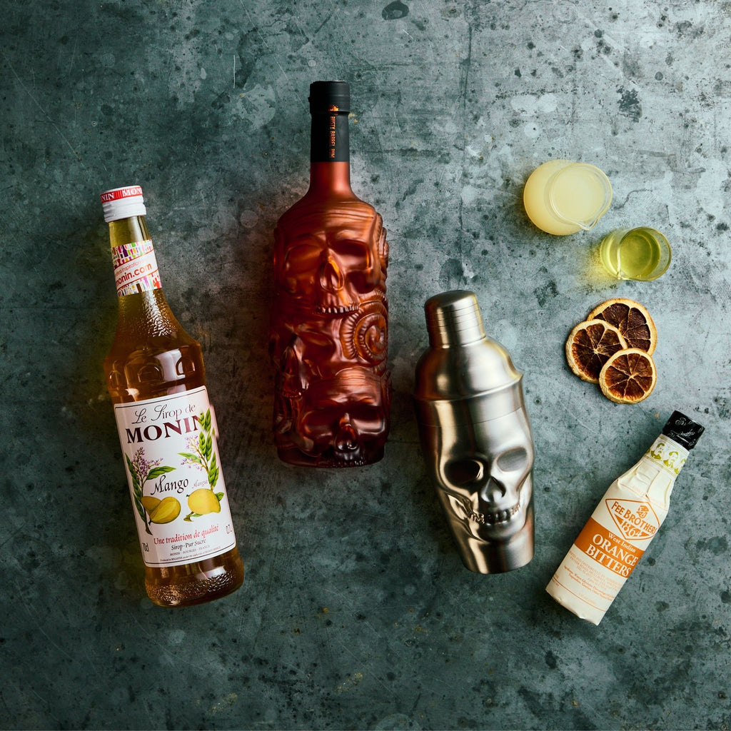 Beverage bottles including Rusty Barrel Gold Rum bottle, a skull shaped cocktail shaker, and a bottle of Monin syrup on a textured surface.
