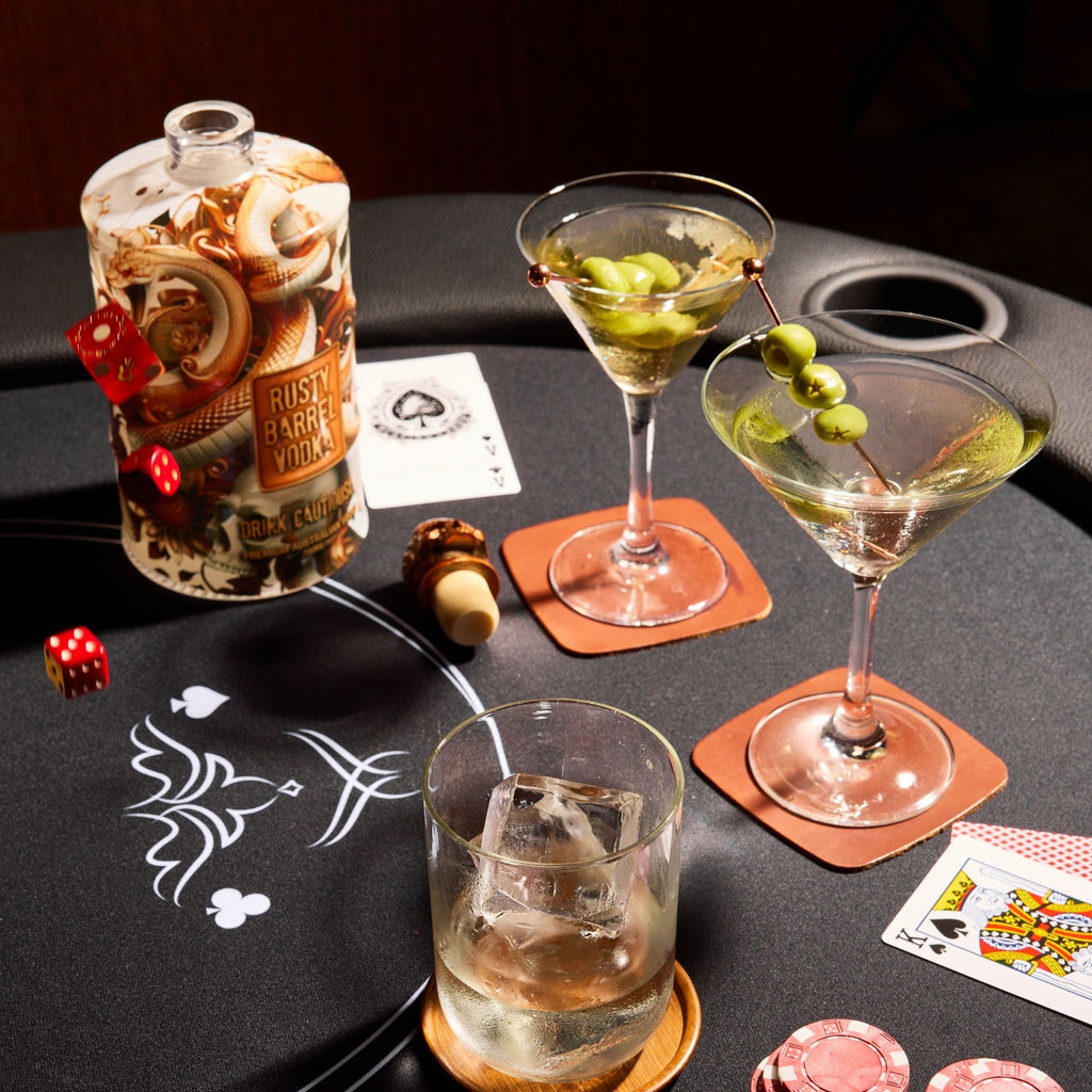 Cocktail glasses with olives, a bottle of Rusty Barre Vodka, dice, and playing cards on a black tray.