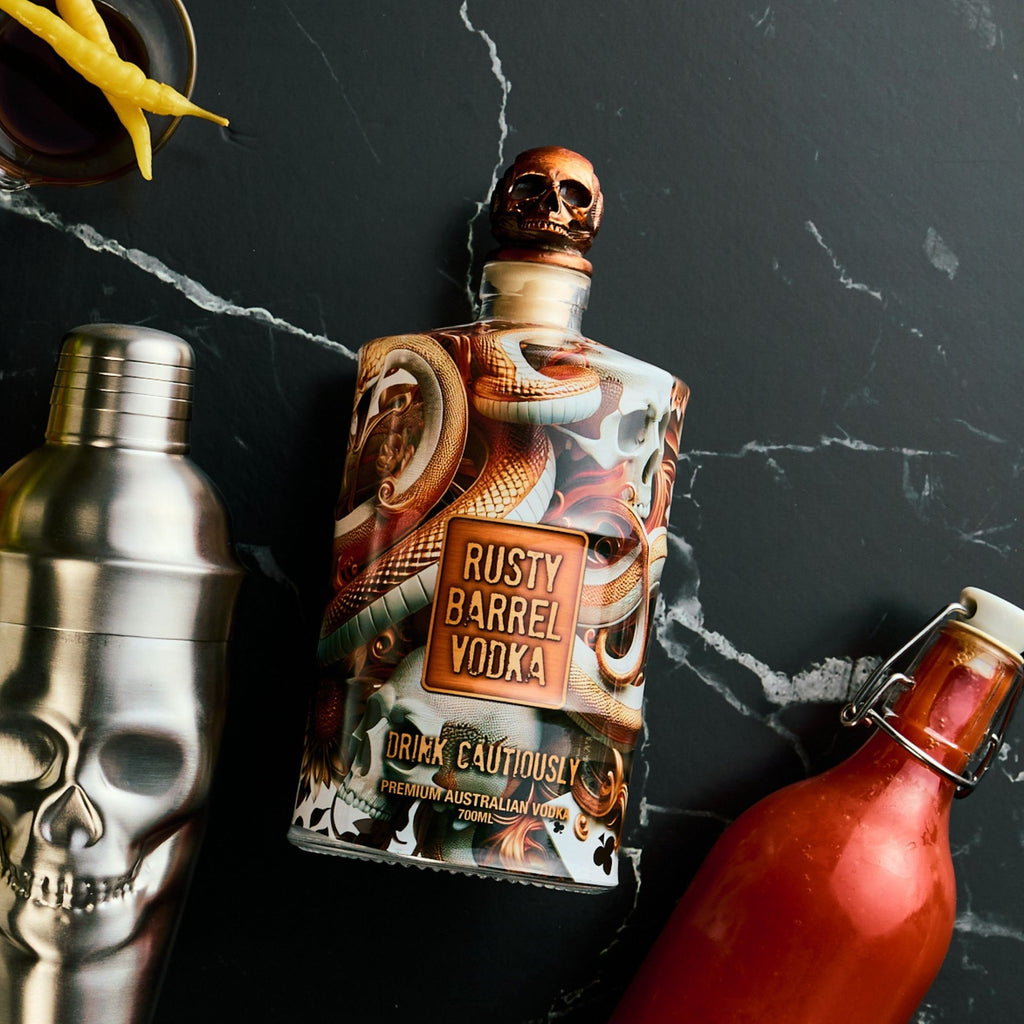 Bottle of Rusty Barrel Vodka with skull design on a dark surface with cocktail shaker and bottle.