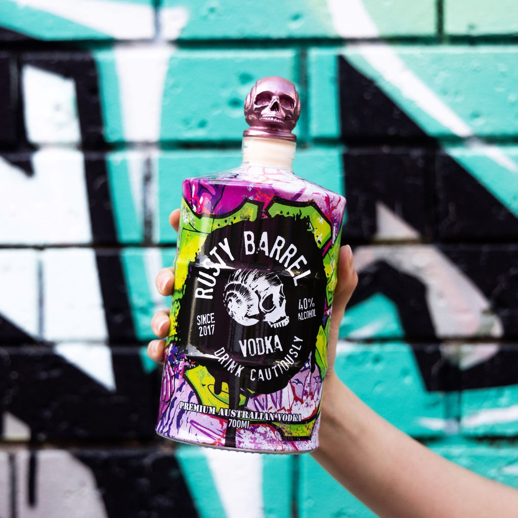 Rusty Barrel Vodka 700ml bottle held in front of a graffiti-covered wall