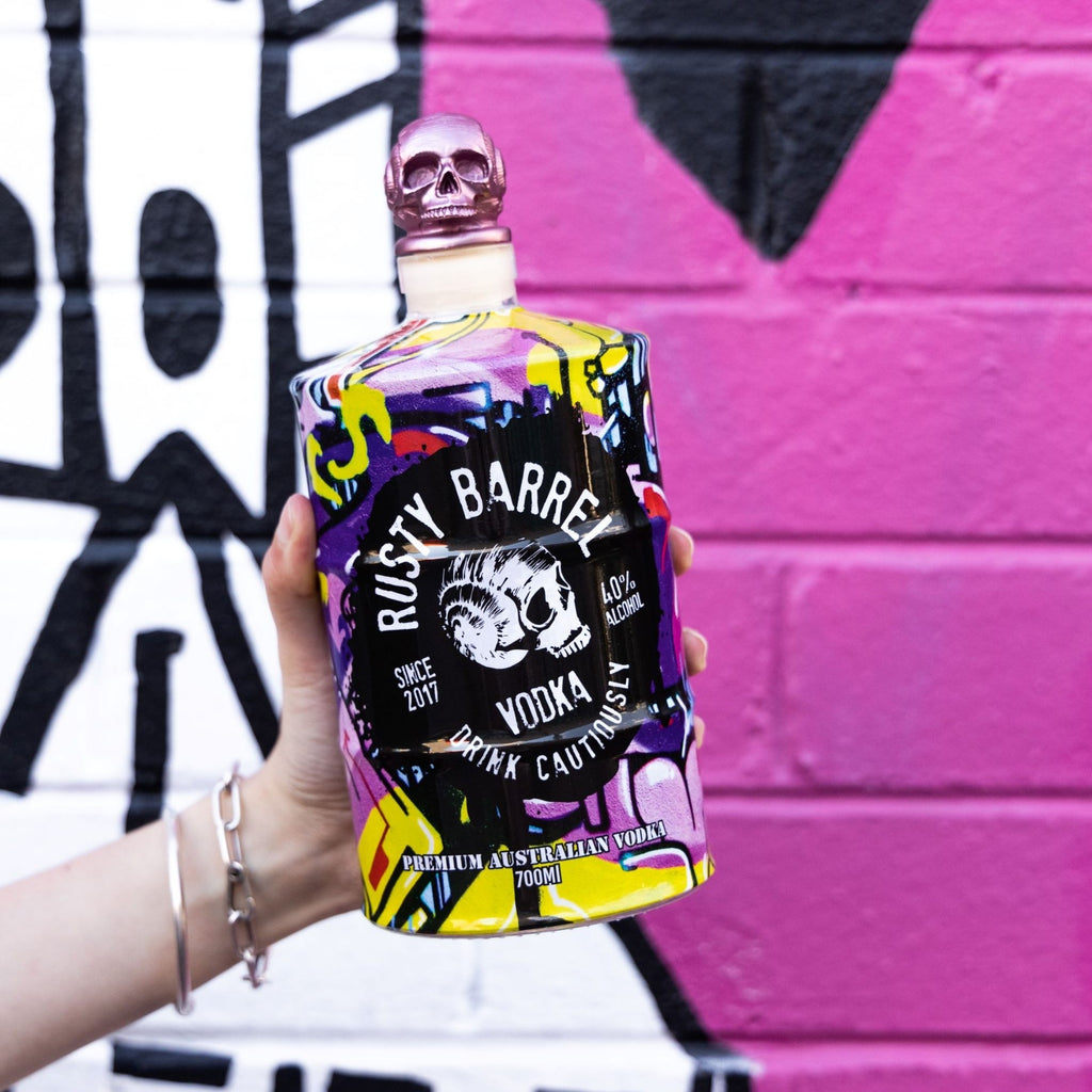 Bottle of Rusty Barrel Vodka 700ml Yellow Wildstyle held in front of a colorful graffiti wall.