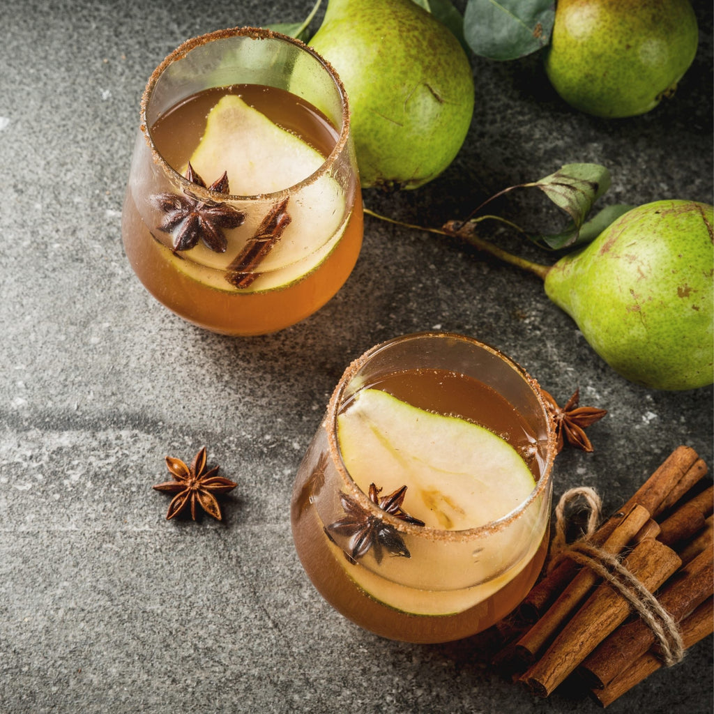 Two glasses of pear-themed cocktails with pears, cinnamon sticks, and star anise on a dark surface.