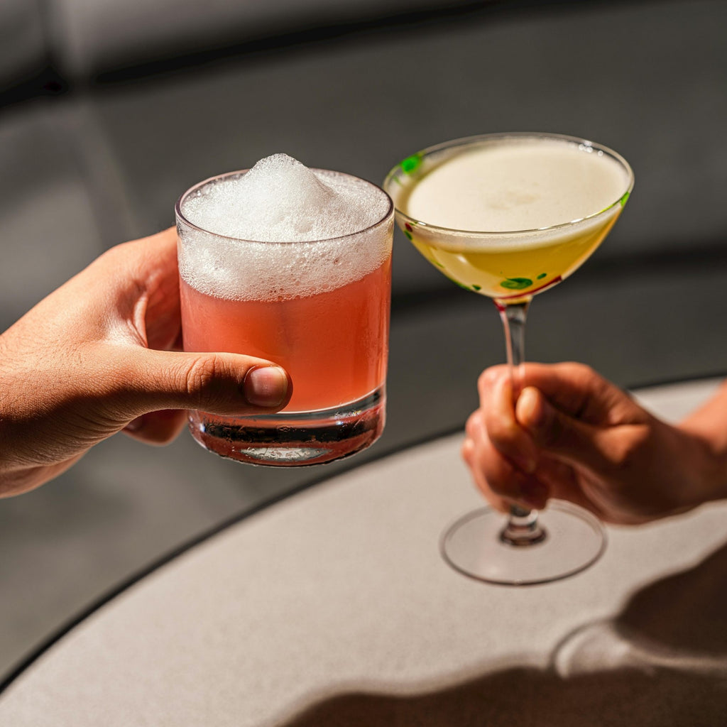 Two hands holding cocktails, one red with foam and the other yellow-green, against a blurred background.